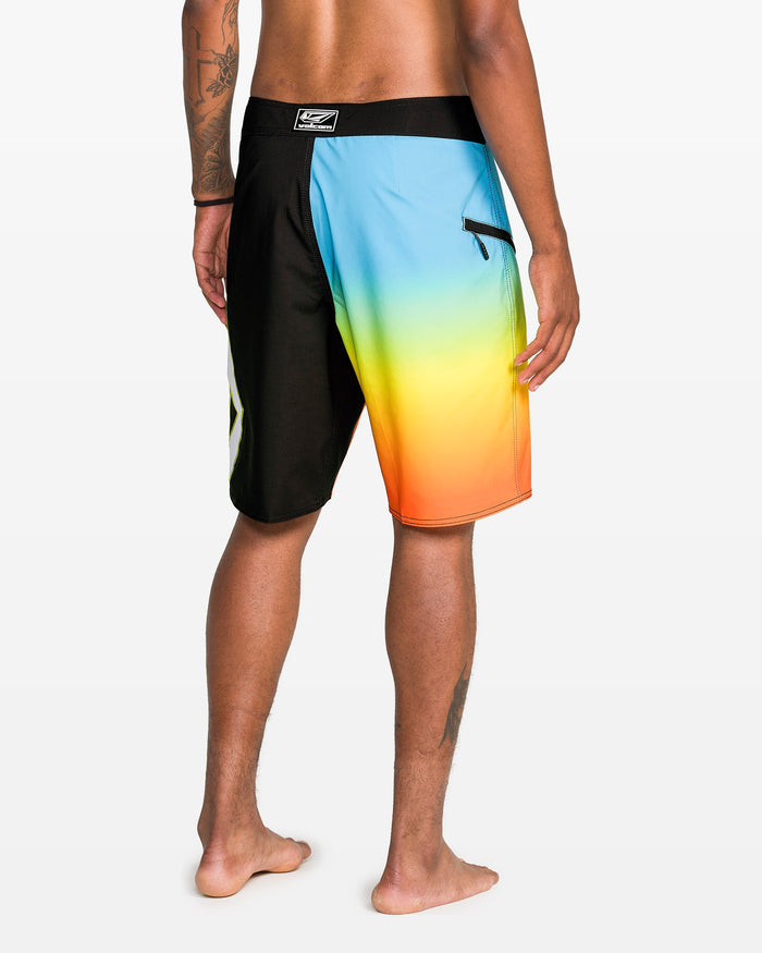 A person stands barefoot, facing away, wearing Volcom Men's Foster Mod 21" boardshorts with a black left side, gradient blue-yellow-orange right side, Cinch Fly Technology, and a zippered pocket.