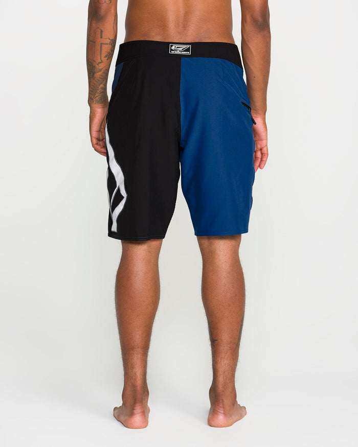 A person seen from behind wears Volcom Men's Foster Mod 21" boardshorts, featuring a loose fit with a half black, half blue color-block design and a wavy white pattern along one side. These boardshorts include a convenient zip side pocket and are worn barefoot against a plain white background.