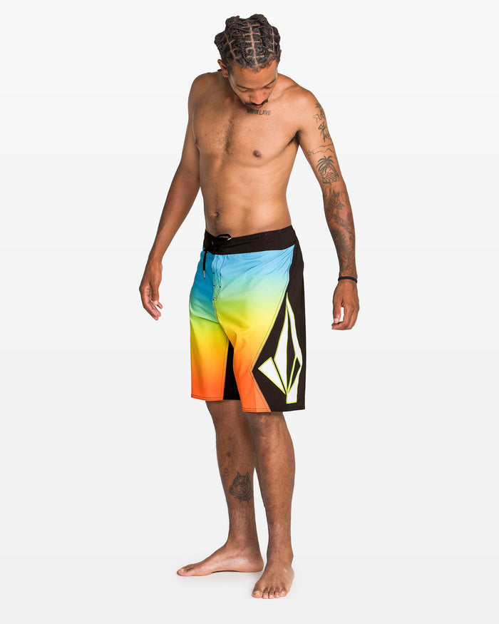 A shirtless, barefoot man with braided hair looks down, wearing Volcom Men's Foster Mod 21" boardshorts. The shorts feature Cinch Fly Technology, a blue-to-yellow-to-orange gradient, and a bold white geometric logo on a plain white background.