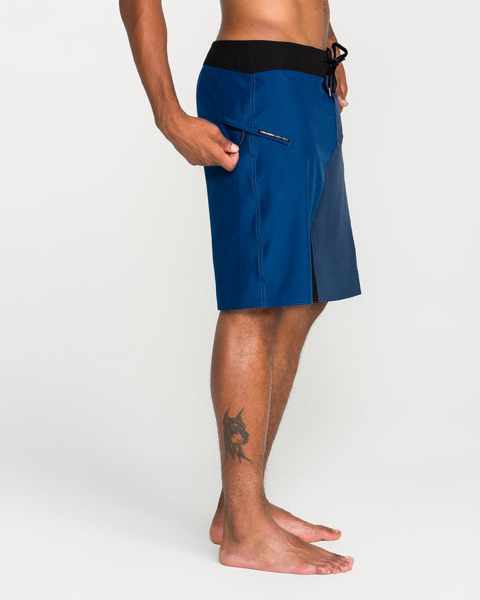 A side profile of a man wearing Volcom Men's Foster Mod 21" boardshorts featuring a blue colorway, black waistband with drawstrings, and a zippered side pocket. The loose fit shorts complement his bare feet, while a dog tattoo on his calf is visible as he poses against a plain light background.