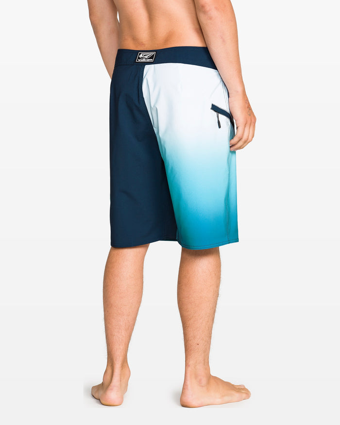 A person stands barefoot facing away, wearing Volcom Men's Foster Mod 21" boardshorts with a blue and white gradient, Cinch Fly Technology, a back zipper pocket, and a 21-inch outseam against a plain white background.