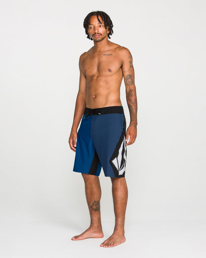 A man with short dreadlocks stands barefoot, wearing Volcom Men's Foster Mod boardshorts—loose fit, 21" length, in blue and black with a geometric design and a convenient zip side pocket. He is shirtless, revealing tattoos on his left arm and chest, set against a plain white background.