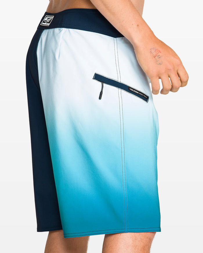 A person is wearing Volcom Men's Foster Mod 21" boardshorts featuring Cinch Fly Technology, a zippered back pocket, and a blue ombre fade from dark navy to light blue. Their left hand with a faded tattoo rests by their side.