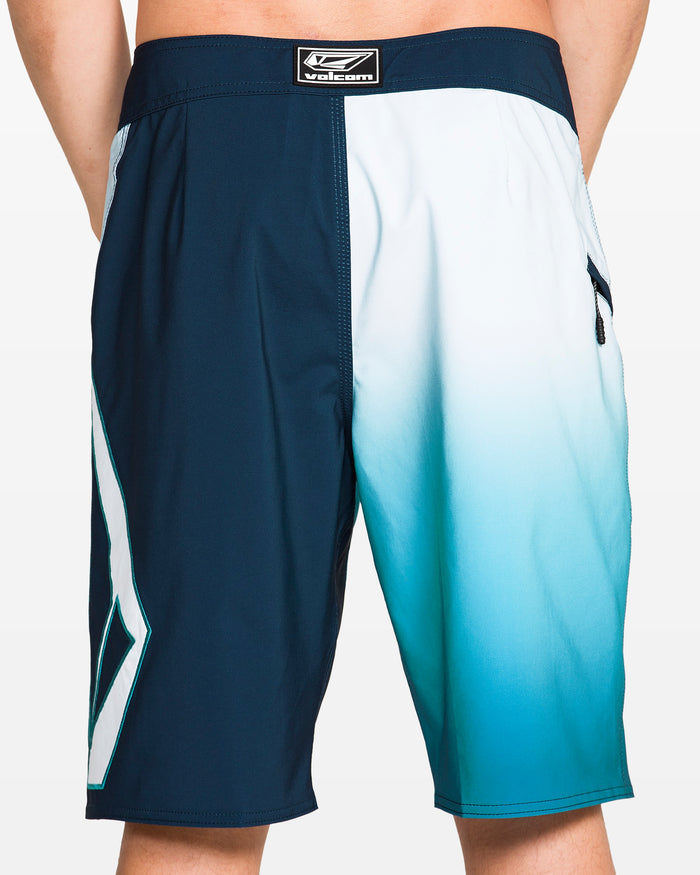 A person stands with hands behind their back wearing Volcom Men's Foster Mod 21" boardshorts featuring a blue and white ombre design, geometric pattern on one leg, Cinch Fly Technology, and a Velcro logo patch on the waistband.