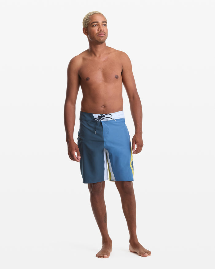 A barefoot man with short blond hair stands against a white background, wearing the Volcom Men's Foster Mod 21 blue board shorts with a black drawstring and white and yellow accents. He is shirtless and looking slightly to the side.