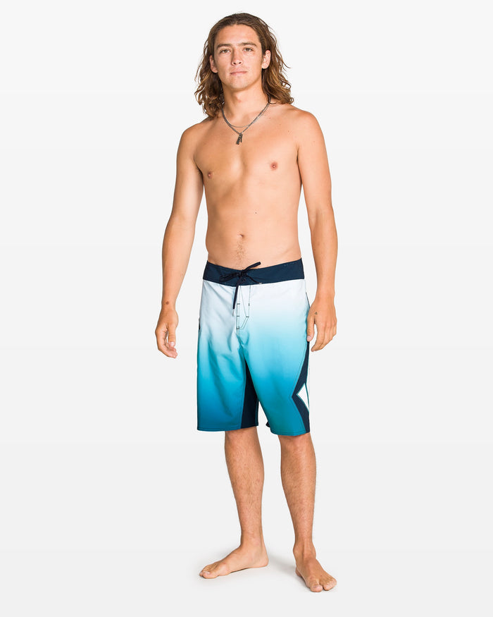 A young man with long hair stands barefoot against a white background, wearing Volcom Men's Foster Mod 21" boardshorts with Cinch Fly Technology. He is shirtless, accessorized with a necklace, and appears relaxed.