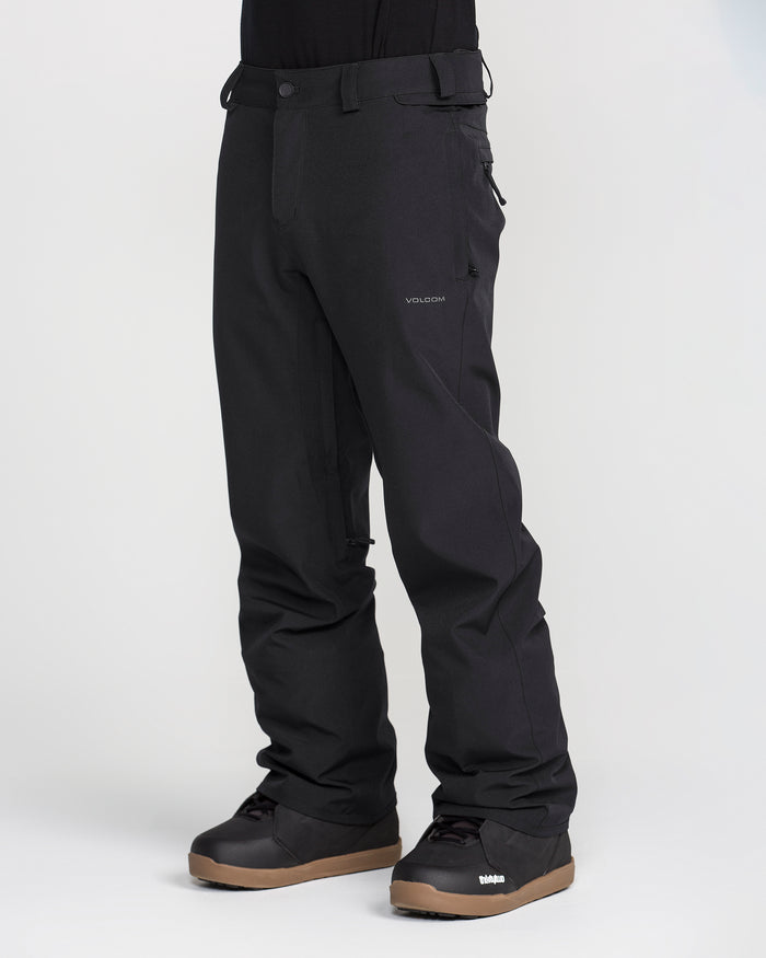 A person wearing black Volcom Men’s Freakin Snow Chino Pants and black winter boots with brown soles stands against a plain light background. The photo focuses on the lower half of the body, accentuating the men’s ski pants crafted from waterproof, breathable fabric by Volcom.