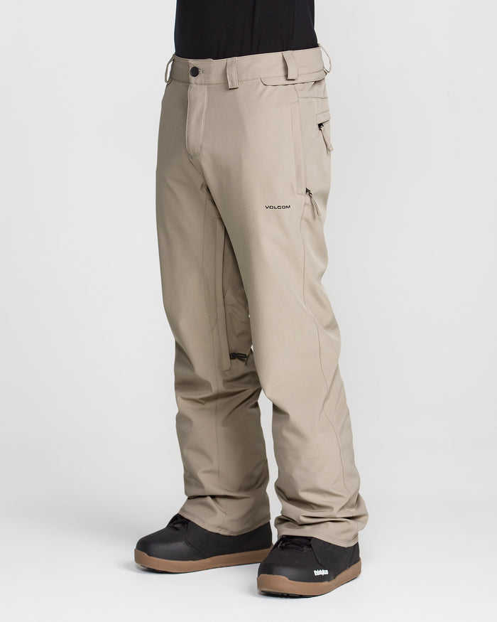 A person is shown from the waist down, wearing Volcom Men’s Freakin Snow Chino Pants in beige—crafted from recycled stretch Oxford fabric—and black snow boots, standing against a plain white background. The waterproof pants have a relaxed fit and zippered pockets.