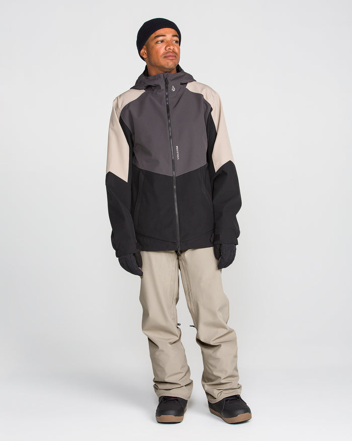 A person stands facing slightly left, wearing a black and gray hooded jacket, Volcom Men’s Freakin Snow Chino Pants in light beige that are waterproof and breathable, black gloves, a black beanie, and dark shoes against a plain white background.