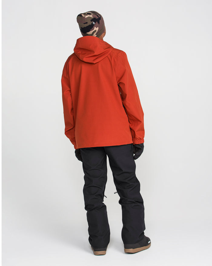 A person wearing a red hooded jacket, Volcom Men’s Freakin Snow Chino Pants in black, black gloves, and a brown camo beanie stands facing away against a plain white background.
