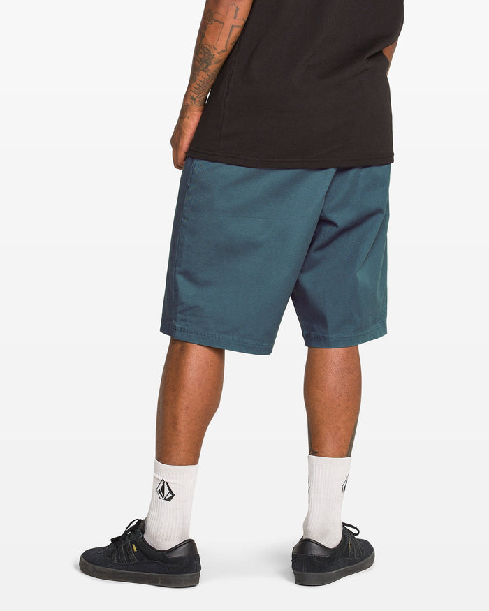 A person stands facing away, wearing a black tee, Volcom Men's Freazy Loose Elastic Waist 22" Shorts, white socks with black logos, and black sneakers. The photo highlights their legs and tattooed arms.