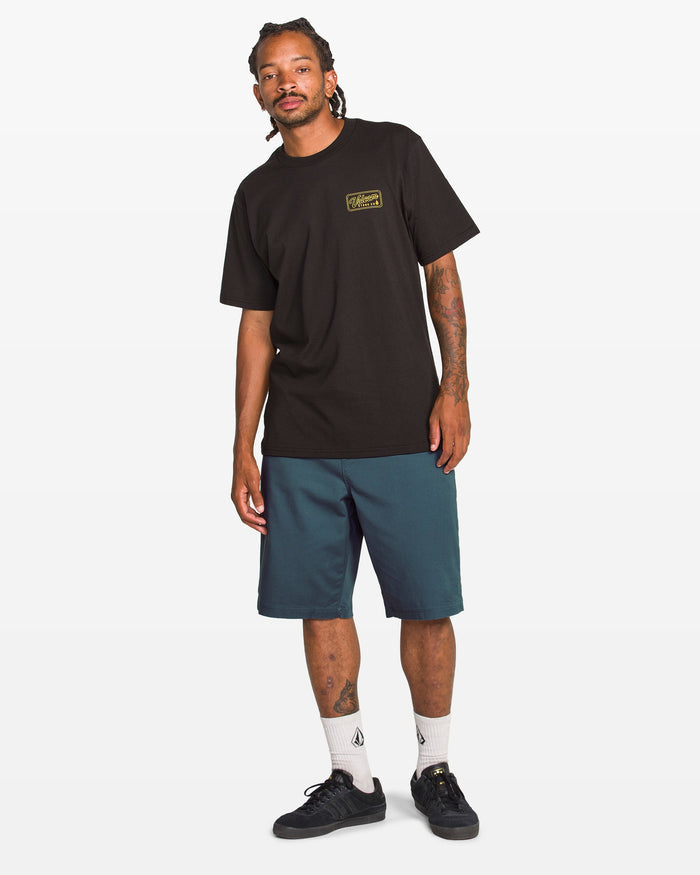 A man stands against a plain white background, wearing a black T-shirt with a small yellow graphic, Volcom Men's Freazy Loose Elastic Waist 22" Shorts in teal, white socks, and black sneakers.