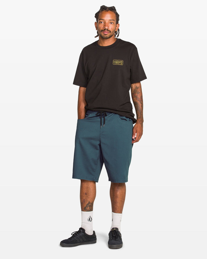A man in casual attire wears a black t-shirt, Volcom Men's Freazy Loose Elastic Waist 22" teal shorts, white socks, and black sneakers against a plain white background.