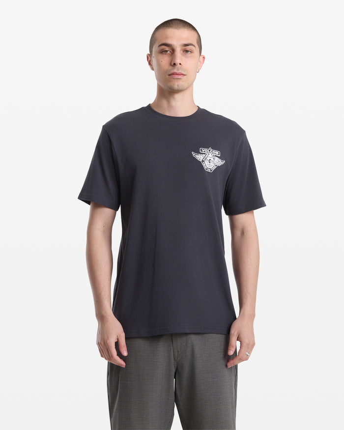 A man with short hair faces forward against a white background, wearing the Volcom Men's Free Wheel Short Sleeve Tee in dark color with a small white left chest graphic and gray pants.