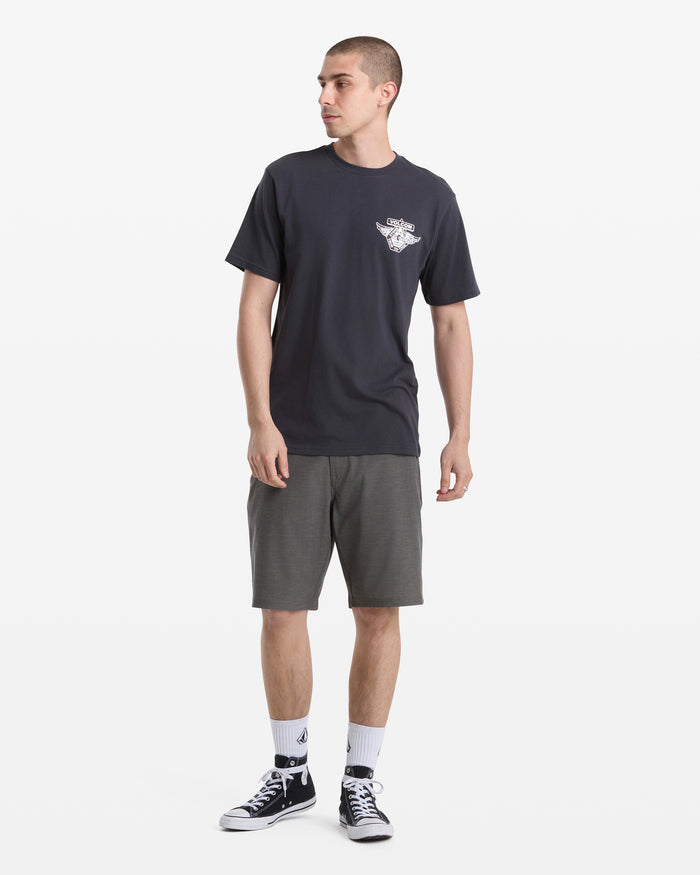 A young man stands against a white background, wearing the Volcom Men's Free Wheel Short Sleeve Tee in black, paired with gray shorts, white socks, and black high-top sneakers. He is looking to his left.