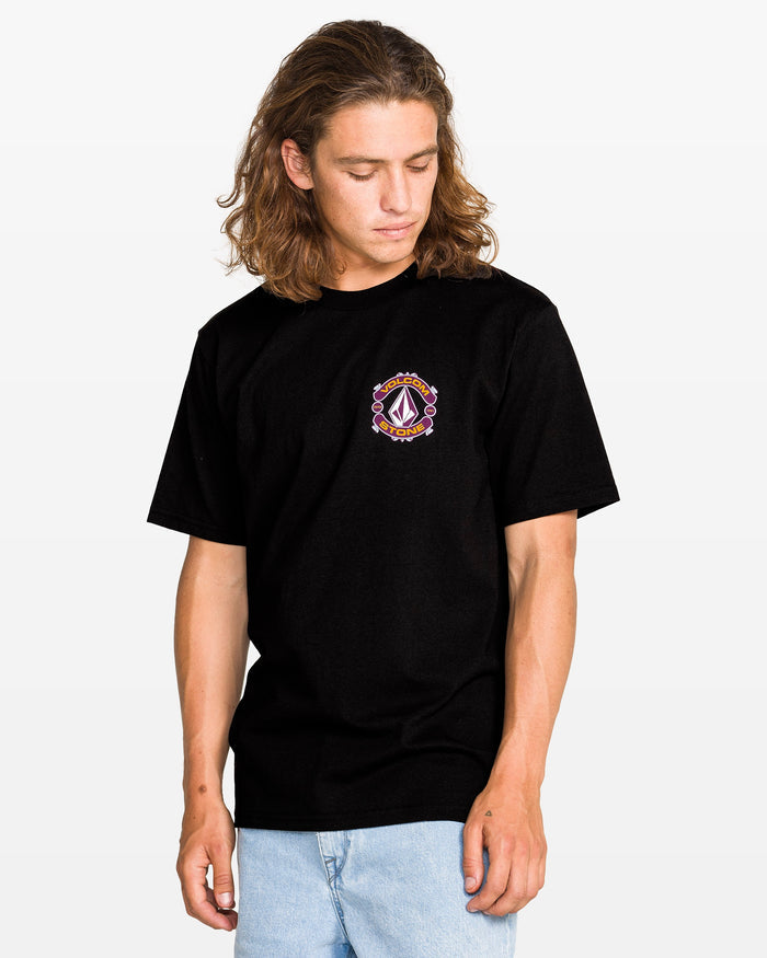 A person with long, wavy brown hair wears the Volcom Men's Freekick Short Sleeve Tee, featuring a colorful small logo on the left chest, paired with light blue jeans, standing and looking down against a plain white background.