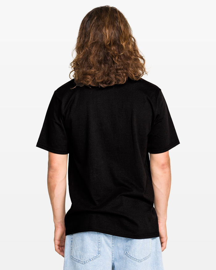 A person with long, wavy brown hair stands facing away, wearing the Volcom Men's Freekick Short Sleeve Tee in breathable cotton and light blue denim shorts against a light background.