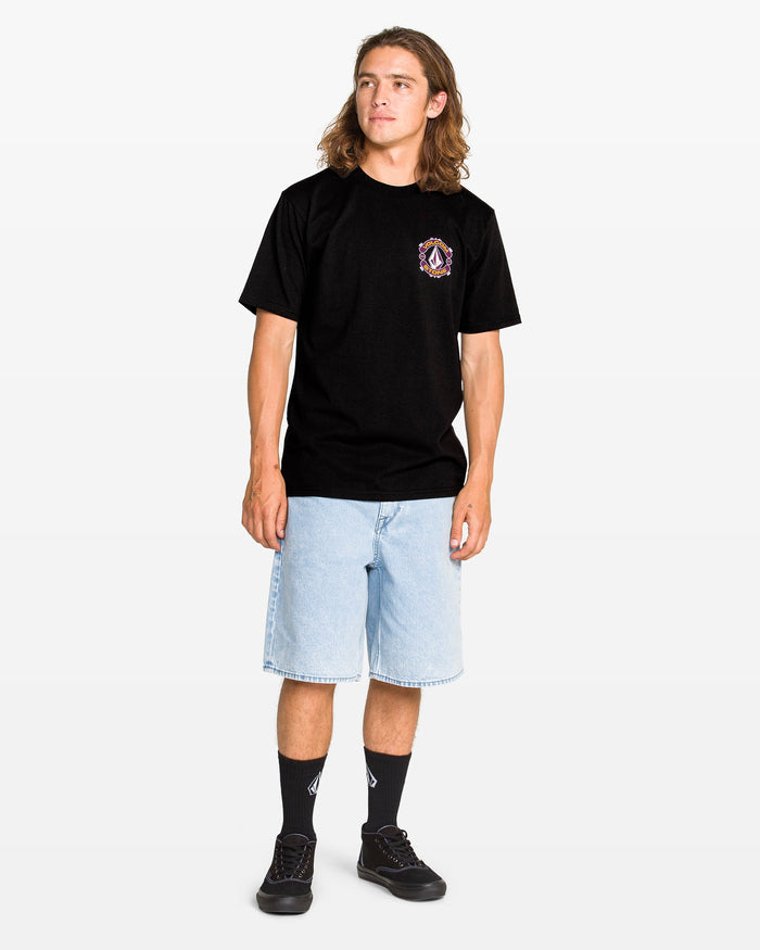 A young man with long hair wears a Volcom Men's Freekick Short Sleeve Tee, light blue knee-length denim shorts, black socks, and black sneakers, standing against a plain white background.