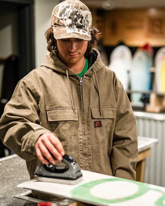 Wearing a tan Volcom Men’s Freshdust Workwear Jacket with a zip front closure and spacious hand and chest pockets, along with a camouflage hat, a person uses an iron on a snowboard in a workshop where surfboards are visible in the background.