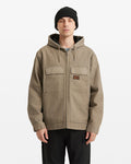 A man in a Volcom Men's Freshdust Workwear Jacket in taupe with large pockets, black pants, and a black knit beanie stands facing forward against a plain white background.