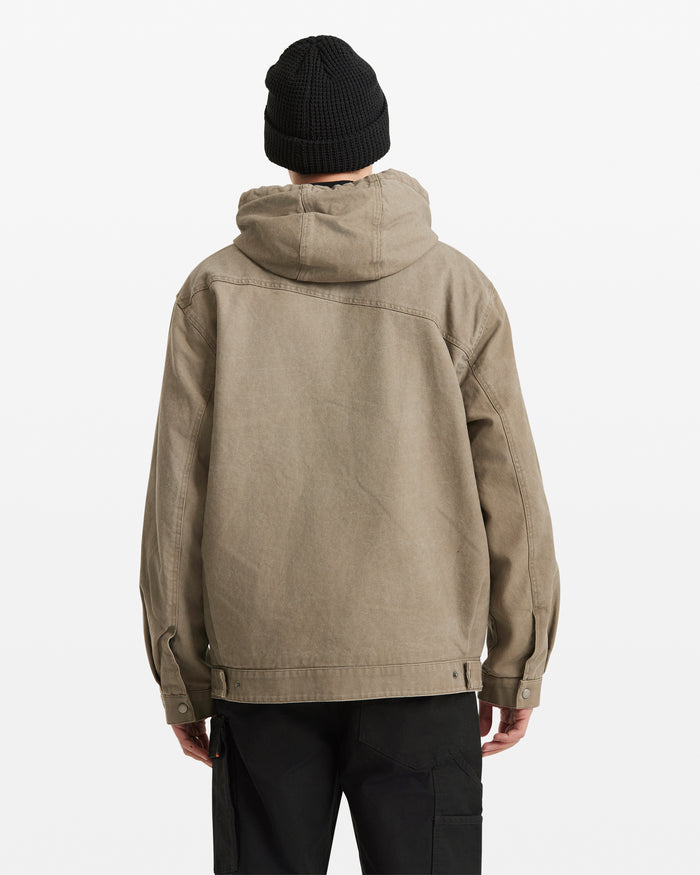 A person wears the Volcom Men's Freshdust Workwear Jacket in tan with a zip front, along with a black beanie and black pants, standing facing away from the camera against a plain white background.