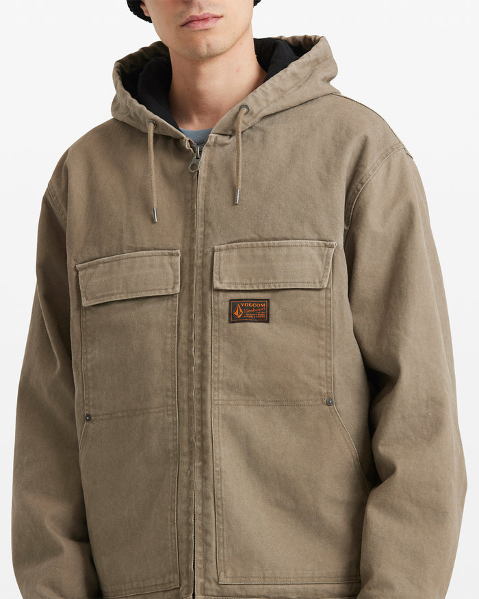 A person wears the Volcom Men's Freshdust Workwear Jacket, featuring large hand and chest pockets, a zip front, and a small rectangular patch on the left chest. Their face is partly visible against a plain background.