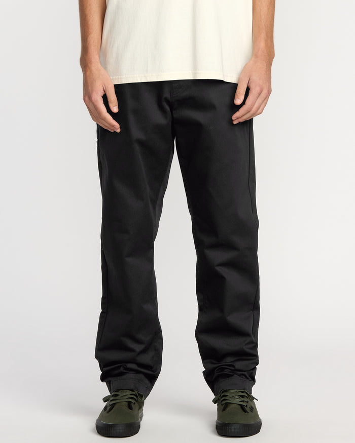 Only the lower torso, legs, and hands are visible as a person stands against a plain background wearing a white t-shirt, Volcom Men's Frickin Chino Pants in Dust Bowl Indigo, and dark green sneakers.