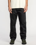 Only the lower torso, legs, and hands are visible as a person stands against a plain background wearing a white t-shirt, Volcom Men's Frickin Chino Pants in Dust Bowl Indigo, and dark green sneakers.