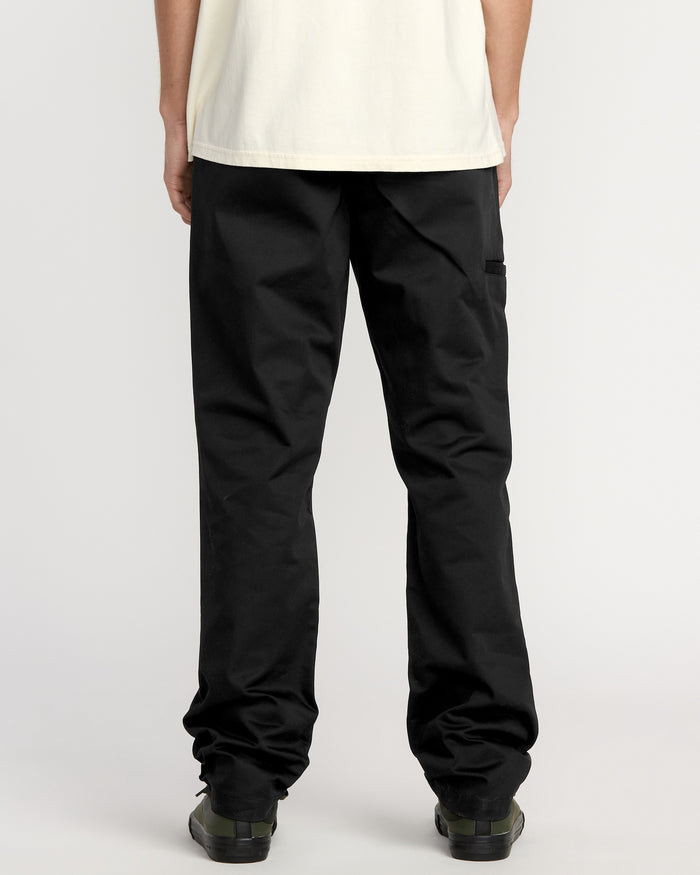 A man faces away from the camera, wearing a cream shirt, black Volcom Men's Frickin Chino Pants (relaxed fit), and black shoes against a plain white background.