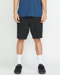 A person stands against a white background wearing a blue shirt, white crew socks, black sneakers, and Volcom Men's Frickin Cross Shred 20" Shorts in black. The image shows the person from shoulders to feet.