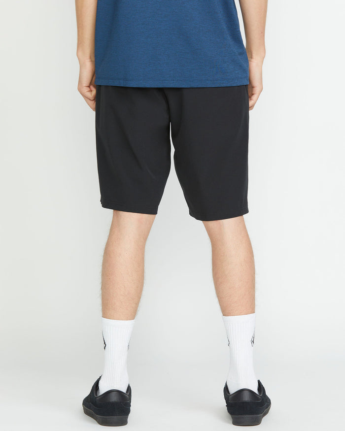 A person faces away from the camera, wearing a blue shirt and Volcom Men's Frickin Cross Shred 20" Shorts in black, paired with white crew socks and black slip-on shoes, set against a plain white background.