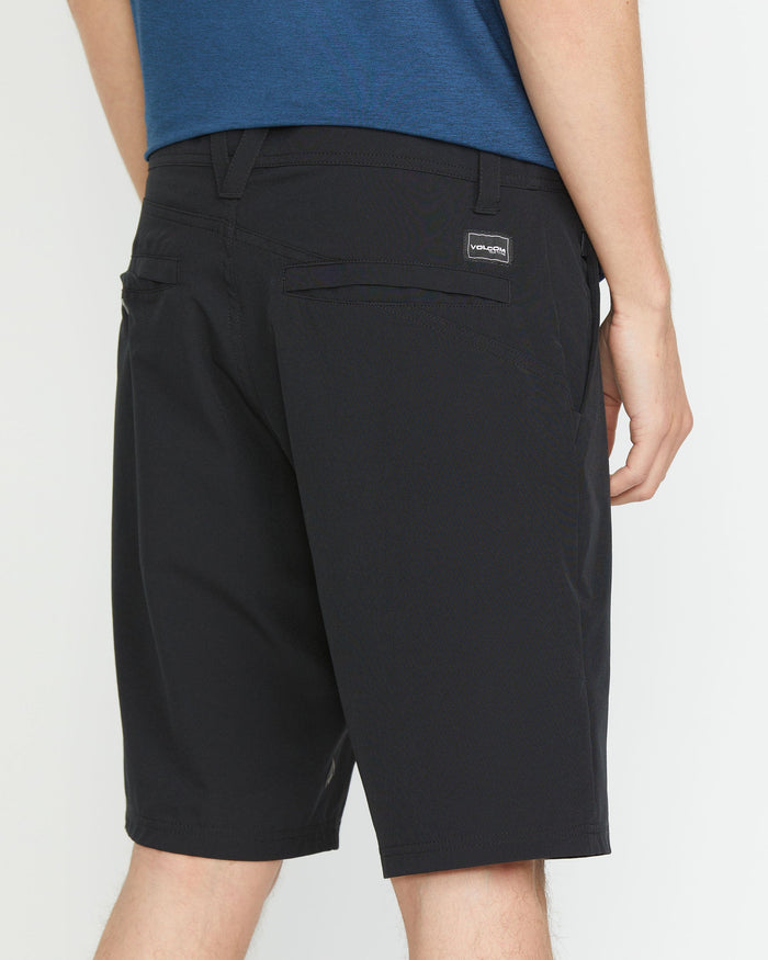 A man wearing a blue shirt and black Volcom Men's Frickin Cross Shred 20" Shorts stands facing away, displaying the shorts' two rear pockets and a small rectangular brand label above the right pocket.