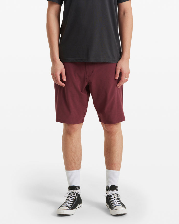 A person wears a black t-shirt, white socks, black Converse sneakers, and Volcom Men's Frickin Cross Shred Static Shorts in maroon. Standing against a plain white background, their head is not visible.