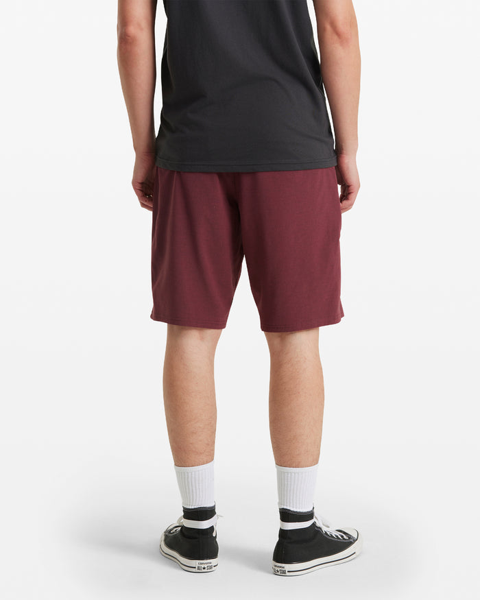A person stands facing away in a black t-shirt, Men's Frickin Cross Shred Static Shorts by Volcom in maroon, white socks, and black low-top sneakers against a plain white background.