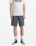 A person, head not visible, stands against a white background wearing blue-gray Volcom Men's Frickin Elastic Waist Shorts, a white graphic T-shirt, white crew socks, and black high-top sneakers.