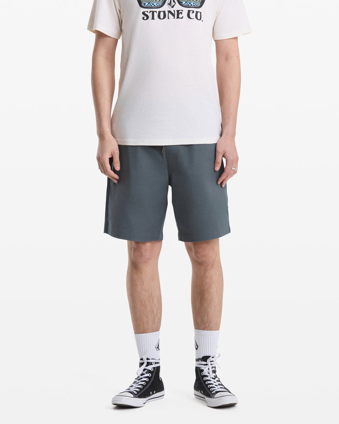 A person, head not visible, stands against a white background wearing blue-gray Volcom Men's Frickin Elastic Waist Shorts, a white graphic T-shirt, white crew socks, and black high-top sneakers.