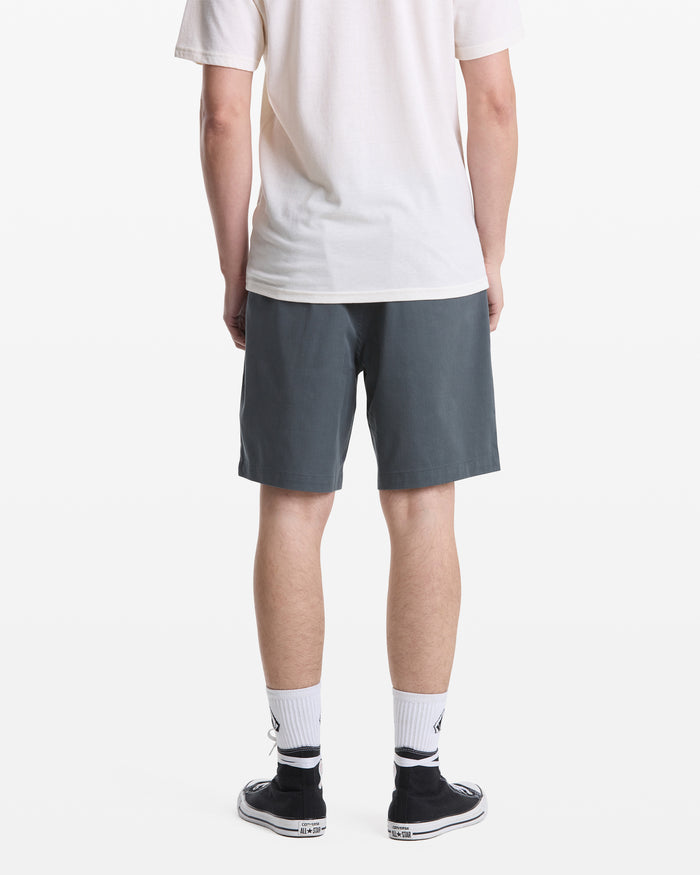 A person stands against a white background, facing away, wearing a white T-shirt, Volcom Men's Frickin Elastic Waist Shorts in dark gray, white crew socks, and black low-top sneakers.