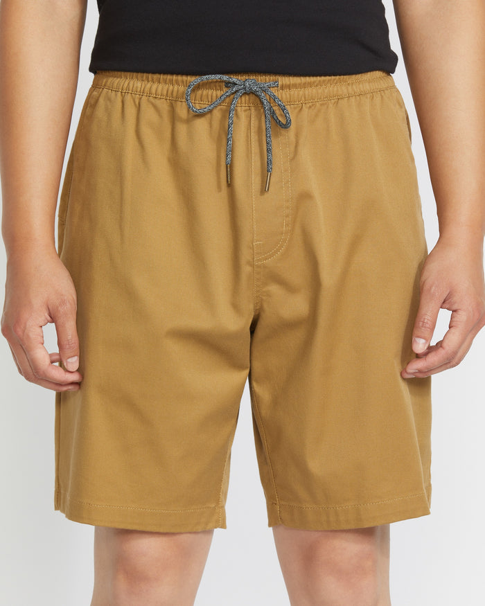 A person wears Volcom Men's Frickin Elastic Waist Shorts in tan, made with REPREVE recycled fibers, and a black top. They stand with arms relaxed at their sides, shown from waist to just above the knees.