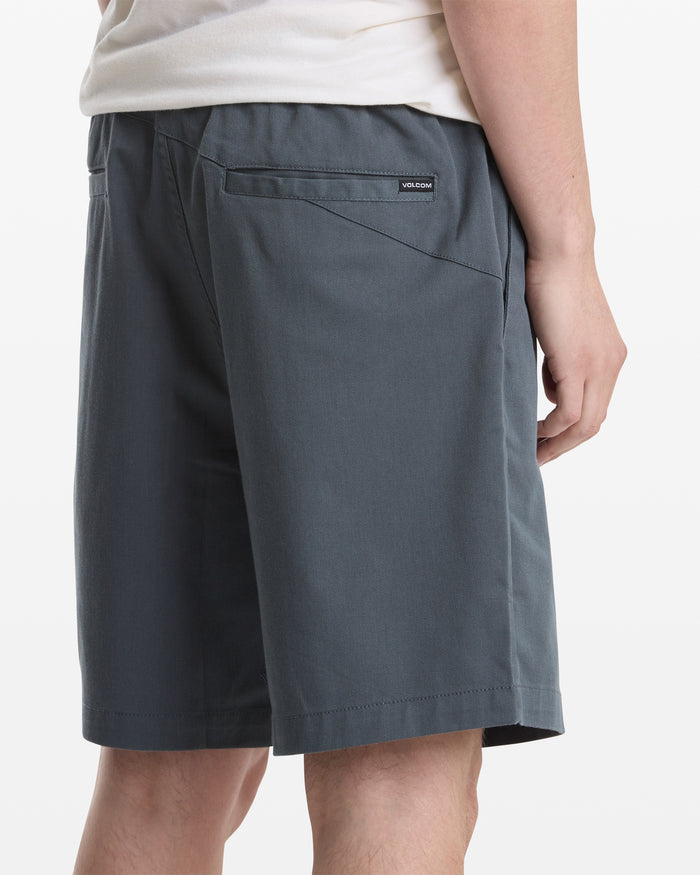 A person wearing a white shirt and Volcom Men's Frickin Elastic Waist Shorts in dark gray is shown from the back and side. The shorts feature a back pocket with a small rectangular Volcom label above it.