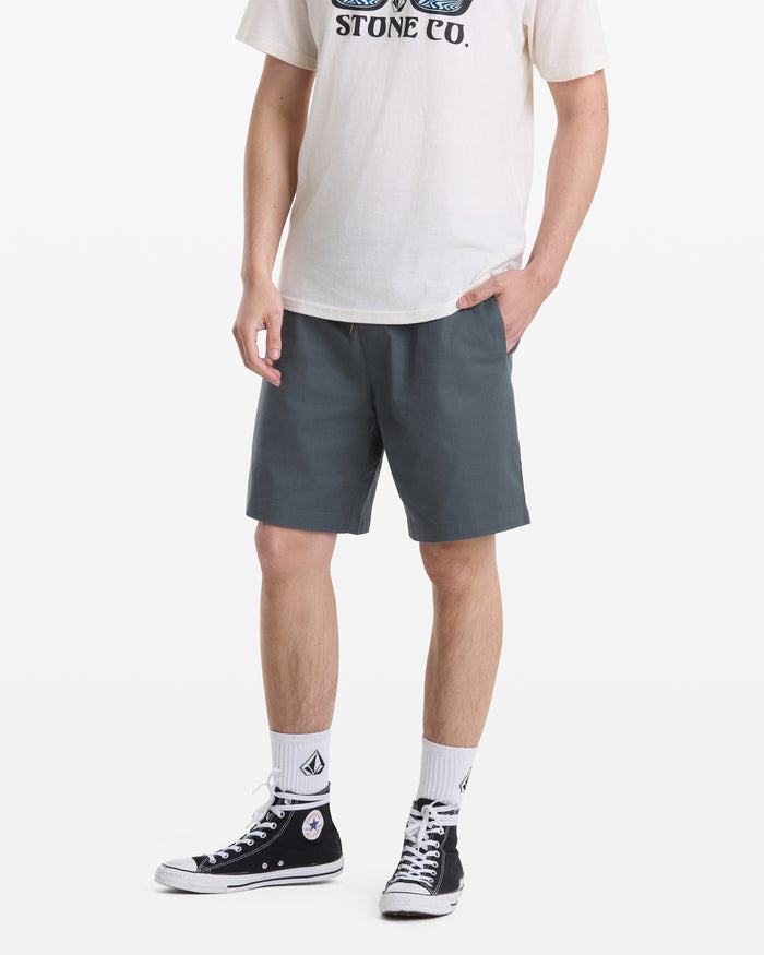 A person stands against a white background wearing a white graphic tee, Volcom Men's Frickin Elastic Waist Shorts in dark gray, white socks, and black high-top sneakers. Their hands are in their pockets; their head is not visible.