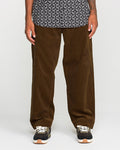 A person showcases the lower half of their body against a simple light backdrop, wearing Volcom’s Men's Frickin Loose Tapered Cord pants in Desert Palm—crafted from 100% cotton corduroy—styled with a black and white patterned shirt and black sneakers featuring white soles.