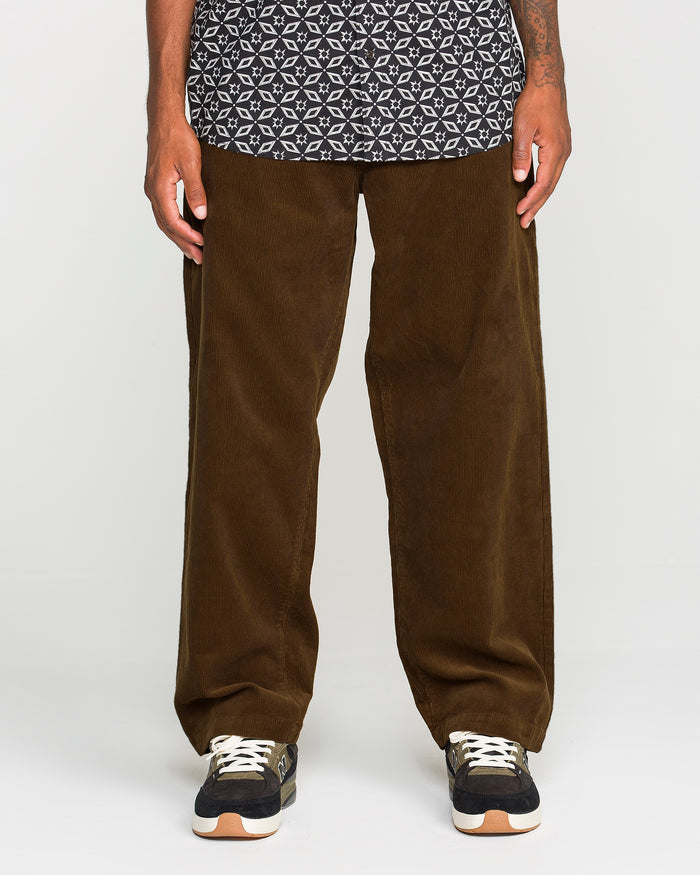 A person showcases the lower half of their body against a simple light backdrop, wearing Volcom’s Men's Frickin Loose Tapered Cord pants in Desert Palm—crafted from 100% cotton corduroy—styled with a black and white patterned shirt and black sneakers featuring white soles.