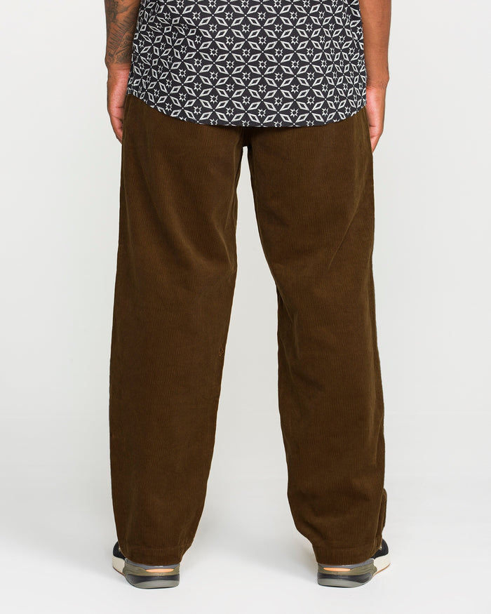 A person stands facing away against a plain light background, wearing Volcom's Men's Frickin Loose Tapered Cord pants in a rich Desert Palm shade. The pants, crafted from 100% cotton corduroy for comfort and style, are paired with a black and white patterned shirt and black shoes featuring white soles.