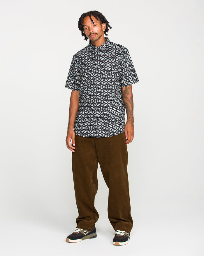 A person stands against a plain background, wearing a black patterned short-sleeve shirt, Volcom Men's Frickin Loose Tapered Cord pants in Desert Palm—a rich hue crafted from 100% cotton corduroy—along with black sneakers. They have short dreadlocks and visible tattoos on one forearm.
