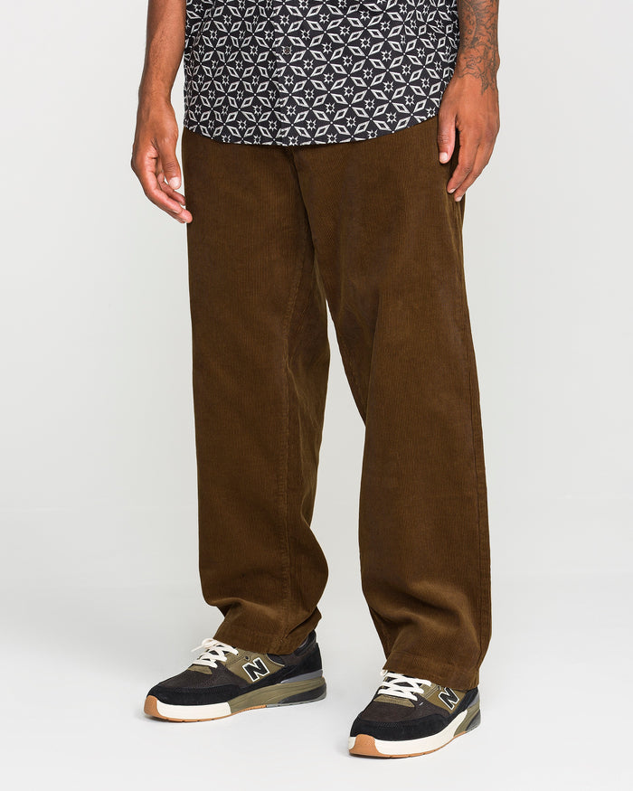 A person wearing a patterned black shirt, the Volcom Men's Frickin Loose Tapered Cord pants in a rich Desert Palm tone made from 100% cotton corduroy, and black New Balance sneakers stands against a plain background.