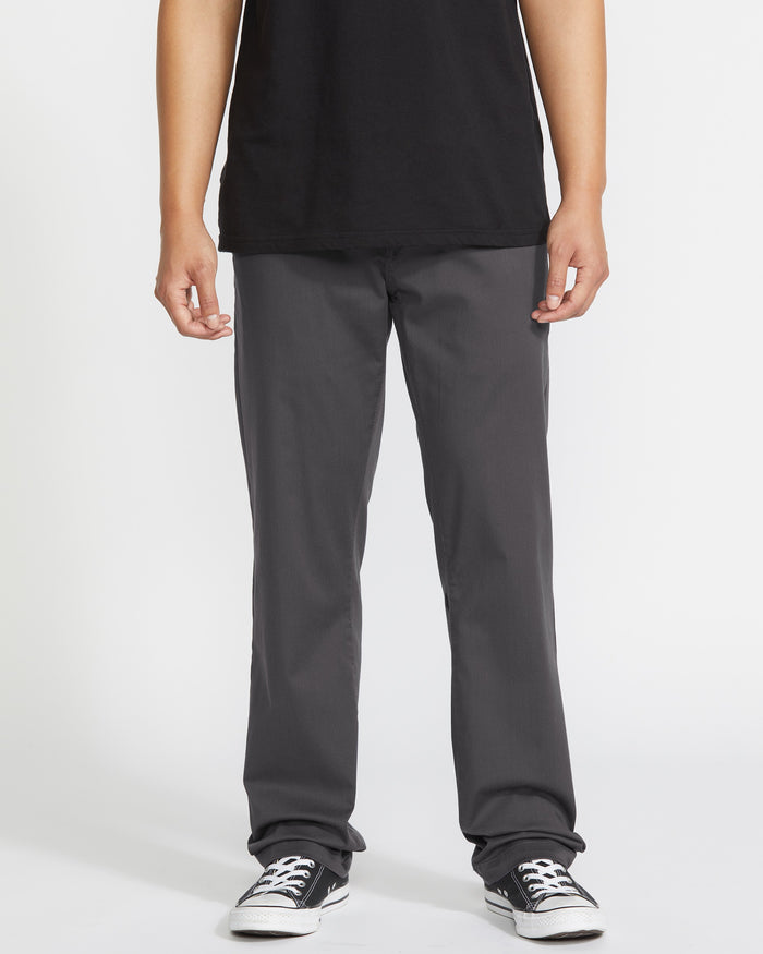 A person wears a black T-shirt, Volcom Men's Frickin Modern Stretch Pants in dark gray, and black-and-white sneakers, standing against a plain white background. The image is cropped at the shoulders.