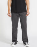 A person wears a black T-shirt, Volcom Men's Frickin Modern Stretch Pants in dark gray, and black-and-white sneakers, standing against a plain white background. The image is cropped at the shoulders.