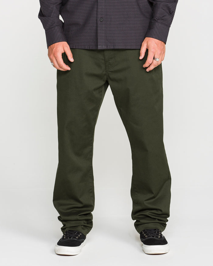 A person wearing Volcom's Men's Frickin Modern Stretch Pants in dark green, crafted with REPREVE recycled fibers, paired with a dark checkered Volcom shirt and black sneakers with white soles, standing against a plain light background.