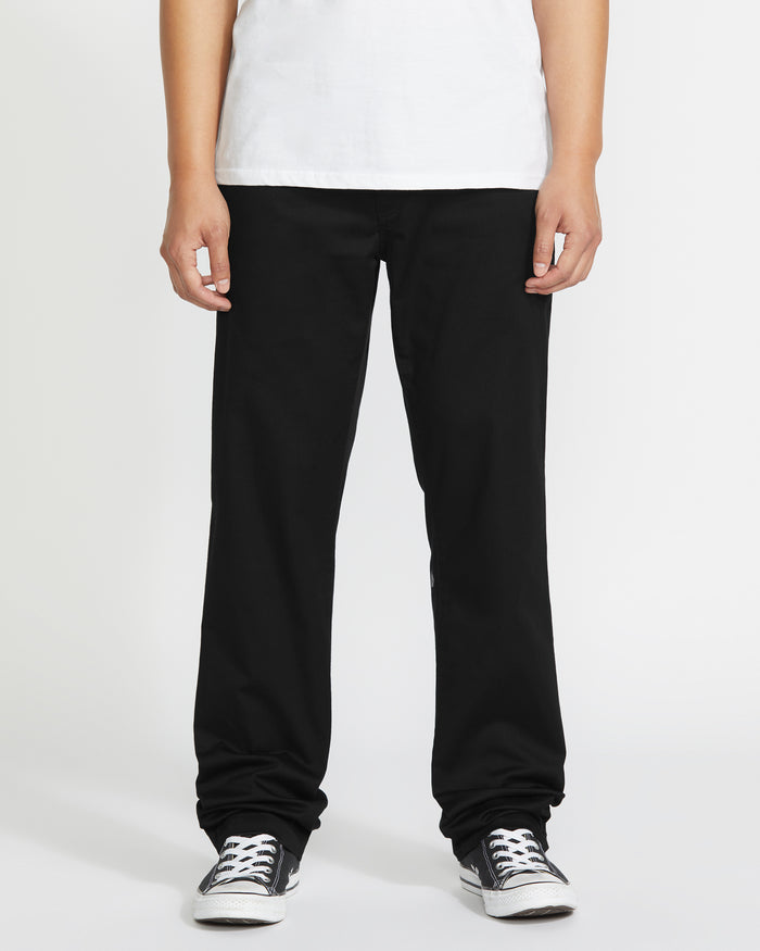 A person stands against a plain background, shown from shoulders down, wearing a white T-shirt, black-and-white sneakers, and Volcom Men's Frickin Modern Stretch Pants in black.