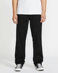 A person stands against a plain background, shown from shoulders down, wearing a white T-shirt, black-and-white sneakers, and Volcom Men's Frickin Modern Stretch Pants in black.